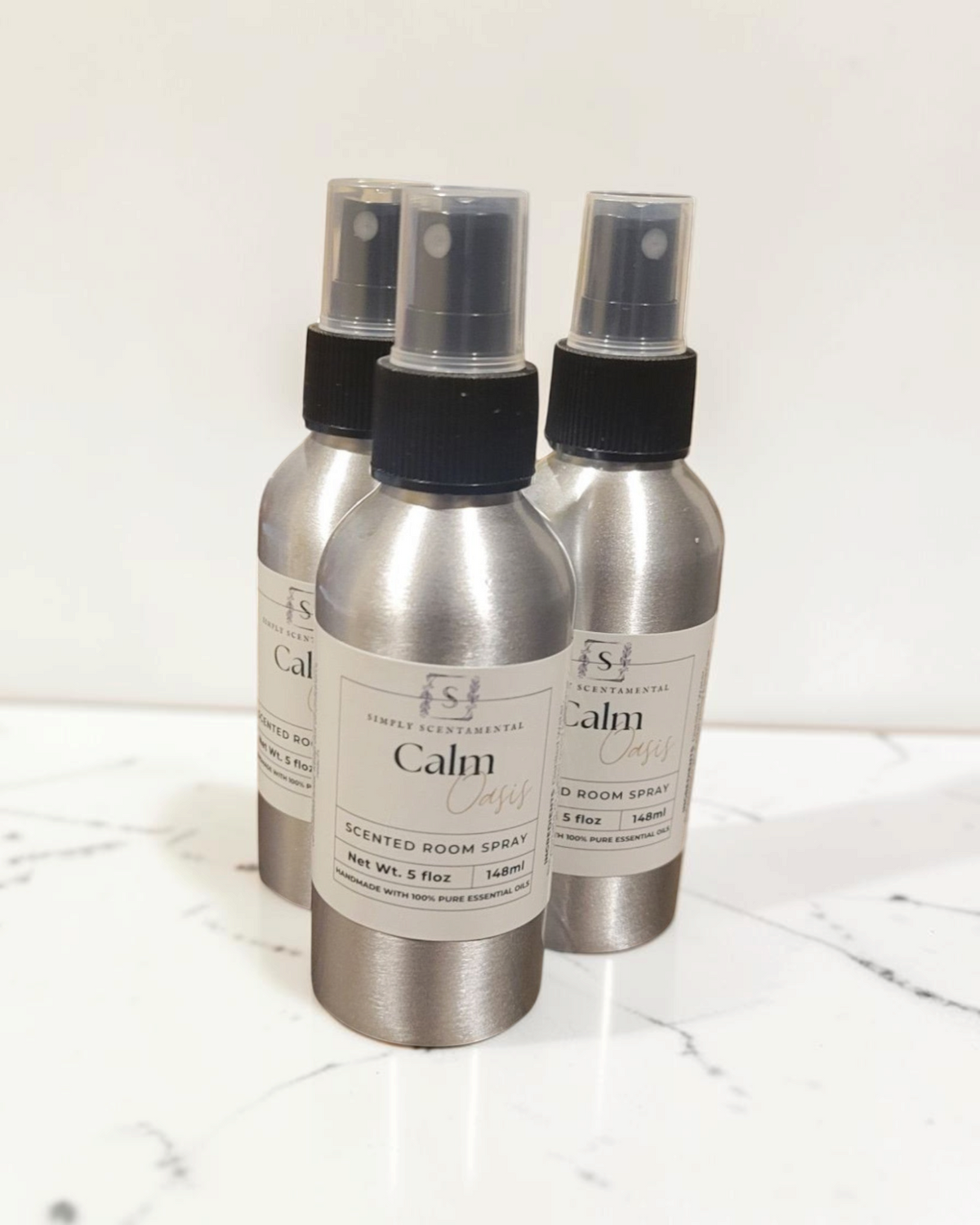 Simply Scentamental Calm Oasis room spray bottles in silver aluminum with black spray nozzles on a wooden surface.